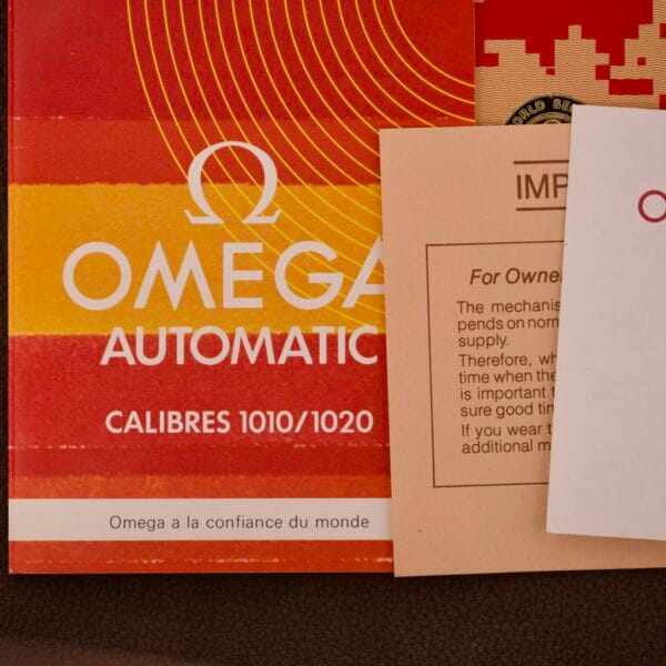 Several Omega watch instruction booklets with bold red and orange designs overlap on a brown background—one highlights the Omega Automatic Seamaster Ref. 166.020 For "U.S. Steel" Cal. 1010 Full Set.