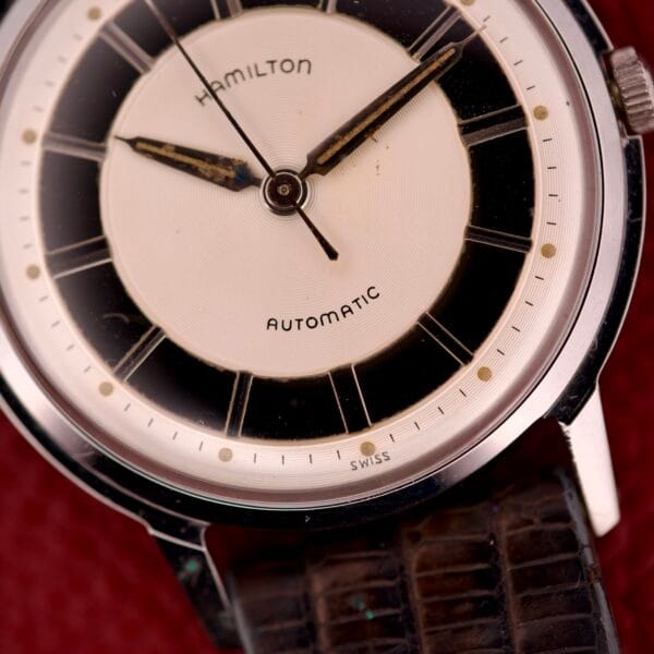 Close-up of a vintage Hamilton Thin-o-matic T-500 wristwatch with Automatic Micro-Rotor Caliber 666 and leather strap on a red textured background.