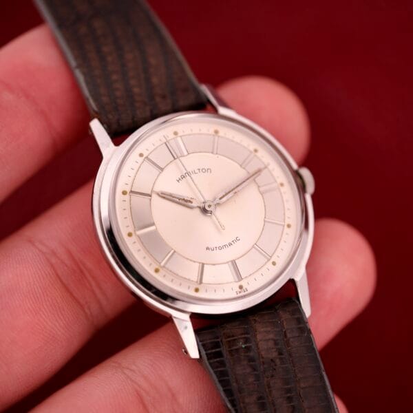 A hand holding a Hamilton Thin-o-matic T-500 Caliber 666 Automatic Micro-Rotor watch with a brown leather strap and cream dial.