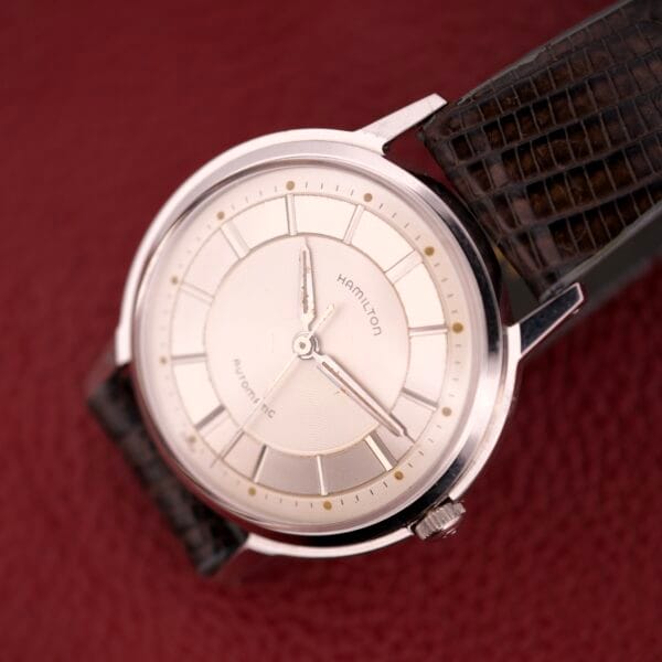 The Hamilton Thin-o-matic T-500 Caliber 666 Automatic Micro-Rotor in silver, featuring a black textured strap, is displayed elegantly on a red textured surface.