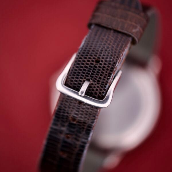 Close-up of a brown textured watch strap with a silver buckle against a red background, ideal for the Hamilton Thin-o-matic T-500 Caliber 666 Automatic Micro-Rotor or other vintage watches of similar style.