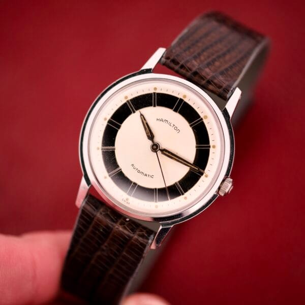 A hand holds the Hamilton Thin-o-matic T-500 Caliber 666 Automatic Micro-Rotor wristwatch, featuring a brown leather strap and a cream-black dial.