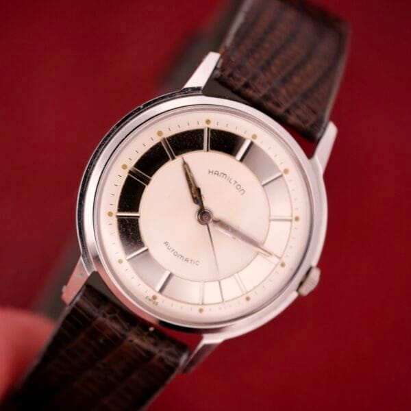 The Hamilton Thin-o-matic T-500 Caliber 666 Automatic Micro-Rotor wristwatch with a brown leather strap and white-black dial is shown against a red background.