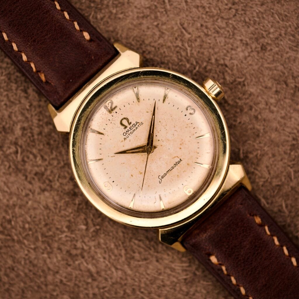 An Omega Automatic Seamaster Ref. 14363 with 14k gold capped bull horn lugs and a brown leather strap rests on a textured brown surface.