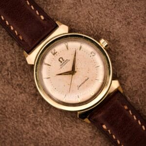 An Omega Automatic Seamaster Ref. 14363 with 14k gold capped bull horn lugs and a brown leather strap rests on a textured brown surface.