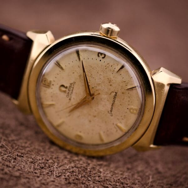 An Omega Automatic Seamaster Ref. 14363 with 14k gold capped bull horn lugs and a brown leather strap rests on a textured surface.