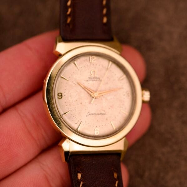 A hand holding an Omega Automatic Seamaster Ref. 14363 with 14k gold-capped bull horn lugs and a brown leather strap.