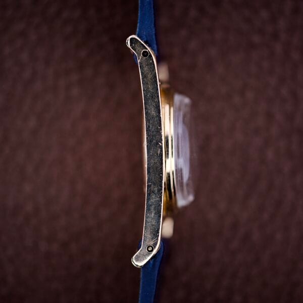 Side view of a Longines Mainliner Art Deco Drivers Watch Manual Wind Cal. 8L, featuring a blue strap and round gold case, displayed against a textured brown background.