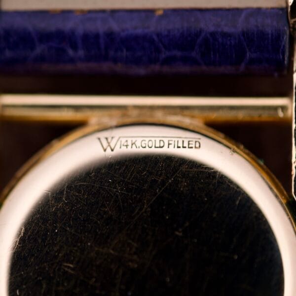 Close-up of a metal surface engraved with W 14K. GOLD FILLED beneath a purple textured edge, from a Longines Mainliner Art Deco Drivers Watch Manual Wind Cal. 8L.