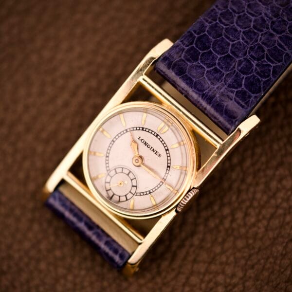 A Longines Mainliner Art Deco Drivers Watch Manual Wind Cal. 8L with a gold rectangular case and textured purple leather strap rests on a brown surface.