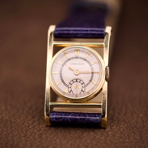 A vintage Longines Mainliner Art Deco Drivers Watch with a manual wind Cal. 8L movement, gold case, and blue leather strap rests on a brown textured surface—where classic design merges with timeless charm.