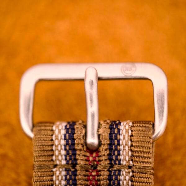 Close-up of a metallic belt buckle with a woven fabric strap, set against a warm orange background inspired by the Nivada Automatic Antarctic Iceberg Date Ref. 4297’s Sunburst Dial.