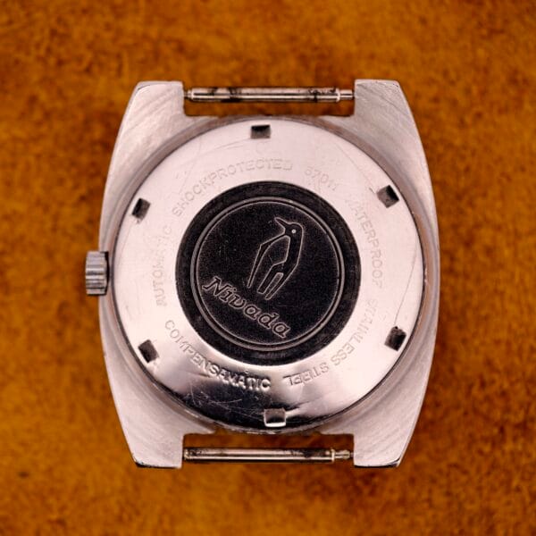 Back of a Nivada Automatic Antarctic Iceberg Date Ref. 4297 Sunburst Dial watch case with penguin logo and engraved text, set on a brown textured surface—a subtle nod to its Antarctic inspiration.