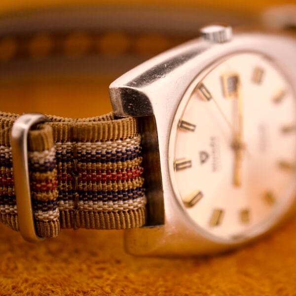 Close-up of the Nivada Automatic Antarctic Iceberg Date Ref. 4297 with sunburst dial, striped fabric strap, and silver case on a yellow textured surface.