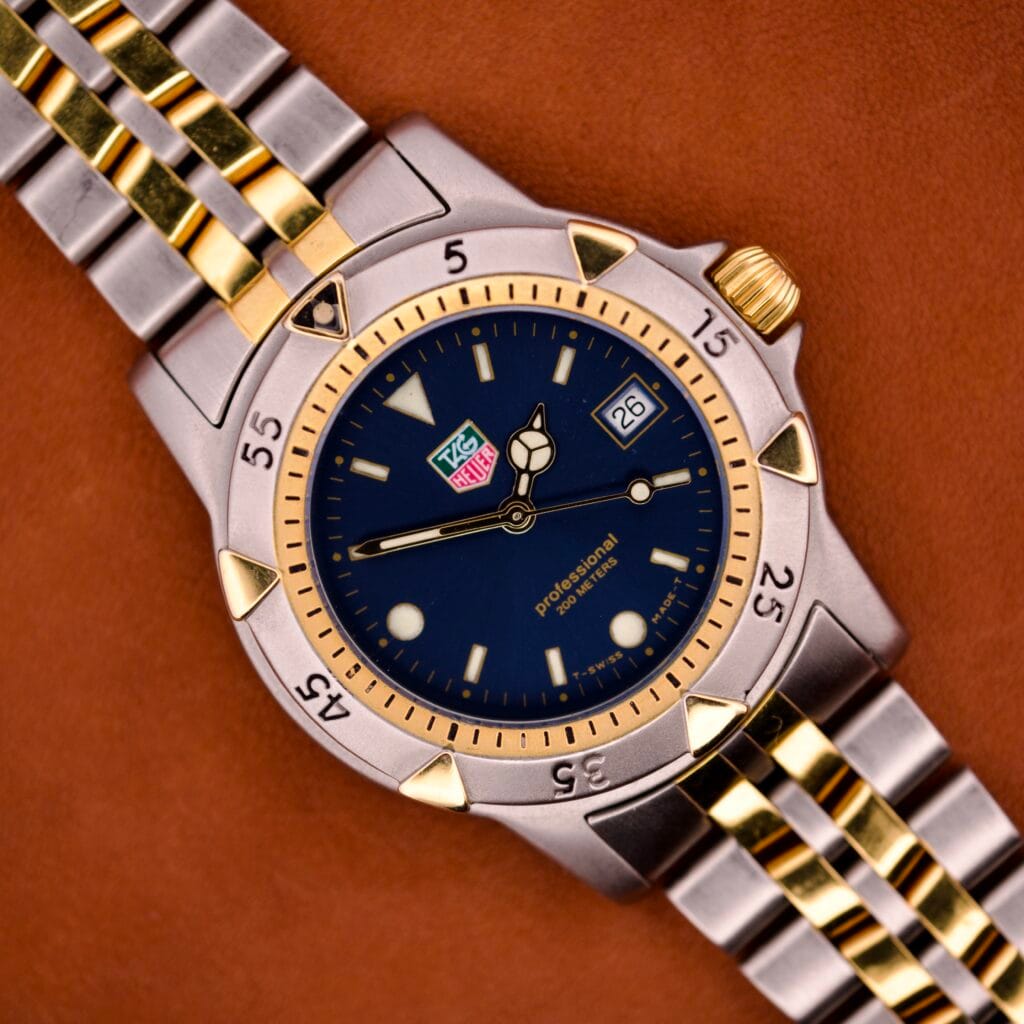 A Tag Heuer 1500 Series Blue Sunburst Dial Ref. WD1221-K-20 wristwatch with date display is shown resting on a brown surface.