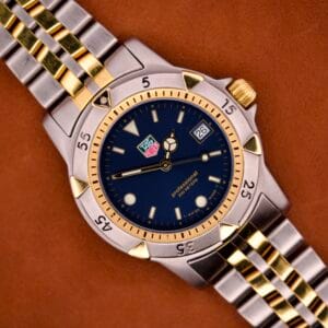 A Tag Heuer 1500 Series Blue Sunburst Dial Ref. WD1221-K-20 wristwatch with date display is shown resting on a brown surface.