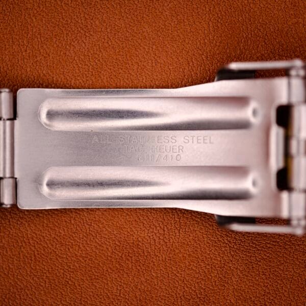 Close-up of a metal clasp engraved with ALL STAINLESS STEEL TAG HEUER 0111/410 on a brown surface, from the Tag Heuer 1500 Series Blue Sunburst Dial Ref. WD1221-K-20—combining timeless design and precision engineering.