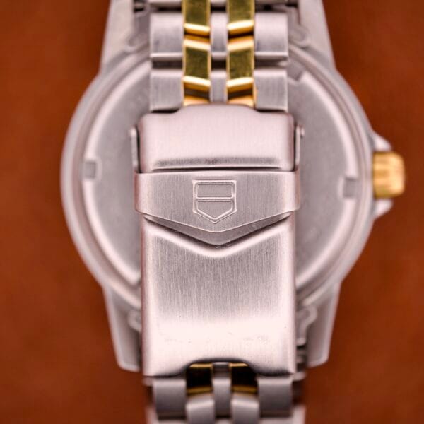 Close-up of a Tag Heuer 1500 Series Blue Sunburst Dial Ref. WD1221-K-20 metal clasp with a brushed finish and gold accents, shown against a brown background.