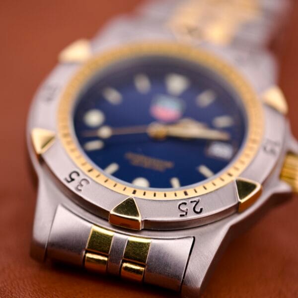 Close-up of a Tag Heuer 1500 Series Blue Sunburst Dial Ref. WD 1221-K-20 wristwatch with gold accents and metal band resting on a brown surface.
