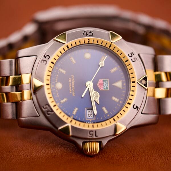 A Tag Heuer 1500 Series Blue Sunburst Dial Ref. WD 1221-K-20 with gold accents and a two-tone metal band is displayed on a brown surface.