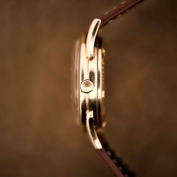 Close-up side view of a Hamilton Automatic Ref. K-407 with a 10k gold filled case and brown leather strap, set against a blurred brown background.