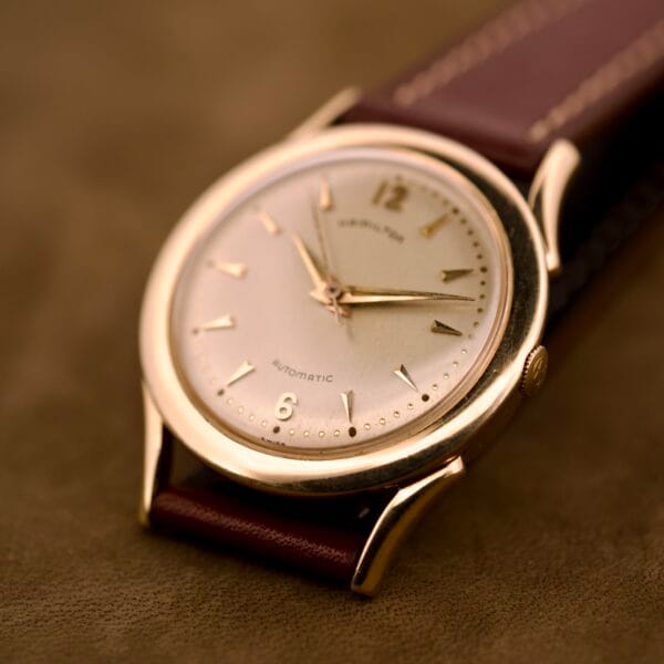 Close-up of a Hamilton Automatic Ref. K-407 wristwatch with a 10k gold filled case and brown leather strap, featuring Cal. 661 movement, displayed on a brown surface.