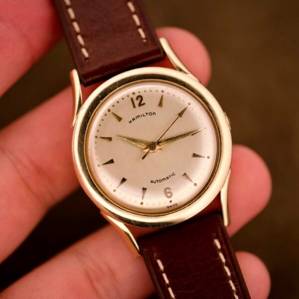 A hand holding a Hamilton Automatic Ref. K-407 wristwatch with Cal. 661 movement, brown leather strap, and 10k gold filled case.
