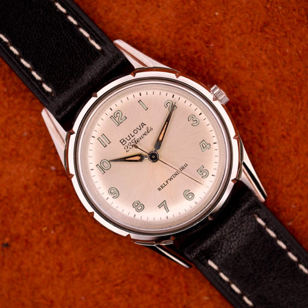 A 1970 Bulova 23 Jewels Self-Winding Cal. 10BZAC watch with a cream dial and black leather strap sits on a textured brown surface.