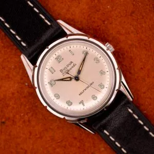 A 1970 Bulova 23 Jewels Self-Winding Cal. 10BZAC watch with a cream dial and black leather strap sits on a textured brown surface.