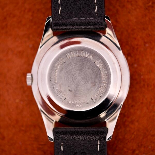 Back of a 1970 Bulova 23 Jewels Self-Winding Cal. 10BZAC wristwatch with engraved text, black leather strap, stainless steel case, and brown background.