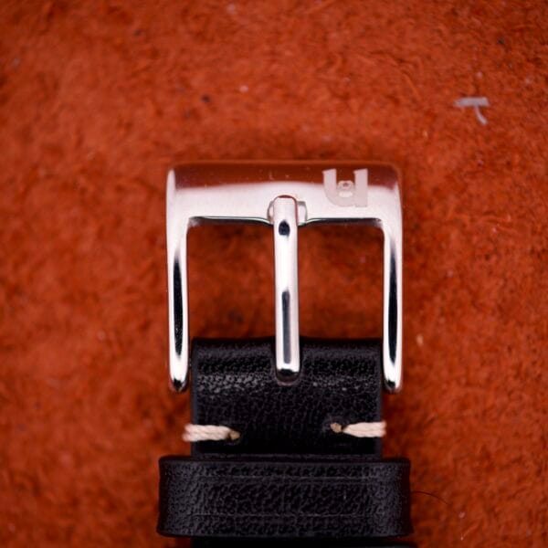 Close-up of a shiny, silver buckle on a black leather belt, evoking the polished elegance of the 1970 Bulova 23 Jewels Self-Winding Cal. 10BZAC stainless steel case, set on a textured reddish-brown background.