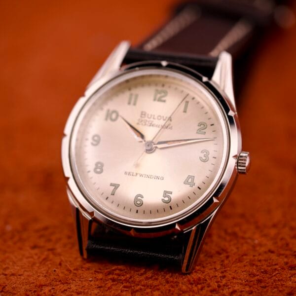 A 1970 Bulova 23 Jewels Self-Winding watch with Cal. 10BZAC movement and stainless steel case, featuring a beige dial and black leather strap, displayed on a brown textured surface.