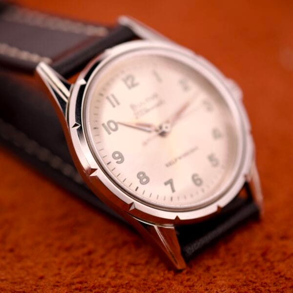 A close-up of the 1970 Bulova 23 Jewels Self-Winding Cal. 10BZAC wristwatch with a cream dial and leather strap on an orange surface, highlighting its classic stainless steel case.