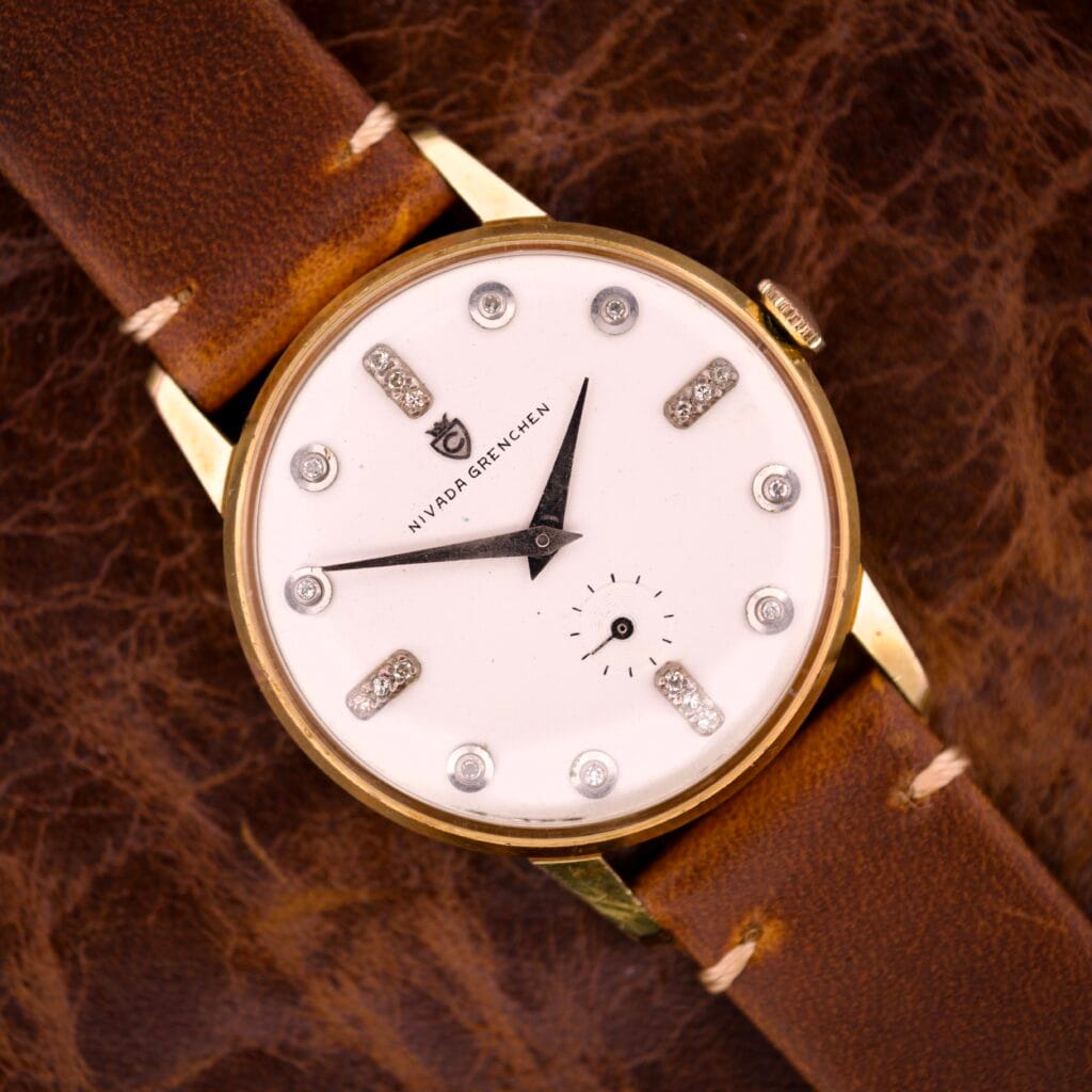 A Nivada Grenchen Manual Wind Cal. R34 with a white dial, diamond markers, and a brown leather strap rests on a textured brown surface.