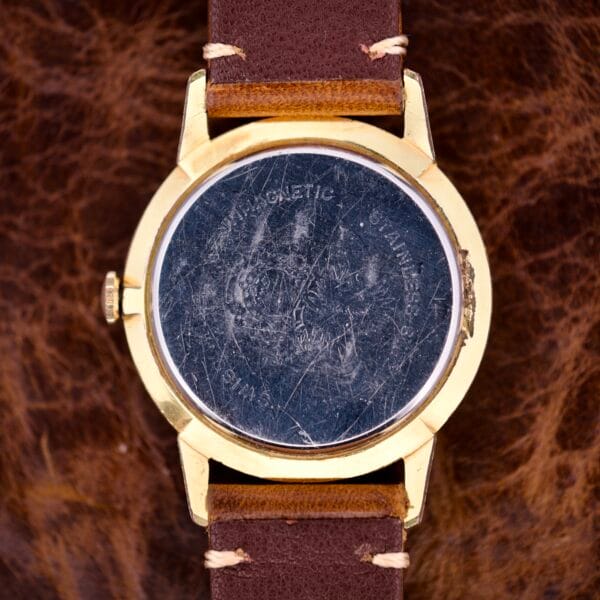 A scratched Nivada Grenchen Manual Wind Cal. R34 with a white dial, diamond markers, and brown leather strap rests on a textured brown surface.