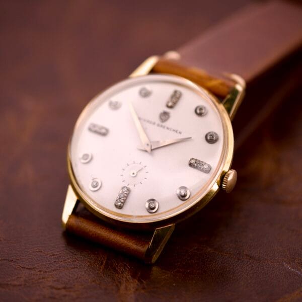 The Nivada Grenchen Manual Wind Cal. R34 features a white dial with diamond markers, a gold case, and a brown leather strap, elegantly displayed on a brown surface.