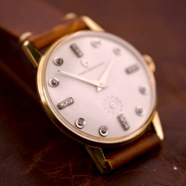 Close-up of a Nivada Grenchen Manual Wind Cal. R34 watch with a white dial, diamond markers, and brown leather strap on a brown surface.