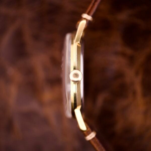 Side view of the Nivada Grenchen Manual Wind Cal. R34 with a white dial, diamond markers, and brown leather strap, set against a blurred brown background.