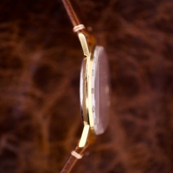 Side view of a Nivada Grenchen Manual Wind Cal. R34 with a white dial, diamond markers, and brown leather strap, set against a blurred brown background.