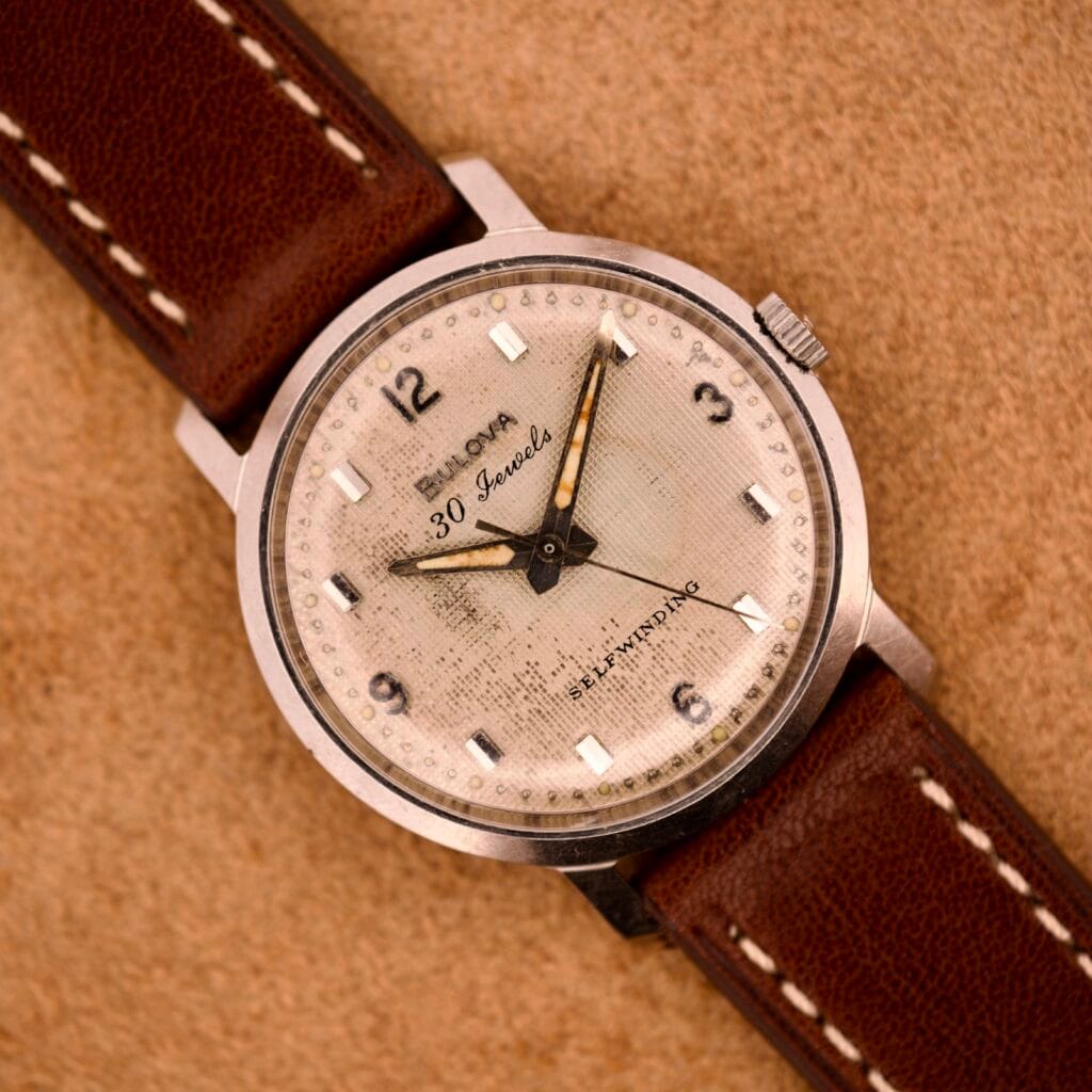 A Bulova 30 Jewels Automatic Cal. 10COAC wristwatch with a stainless steel case and waffle dial rests on a tan surface, set to 10:10 and paired with a brown leather strap.