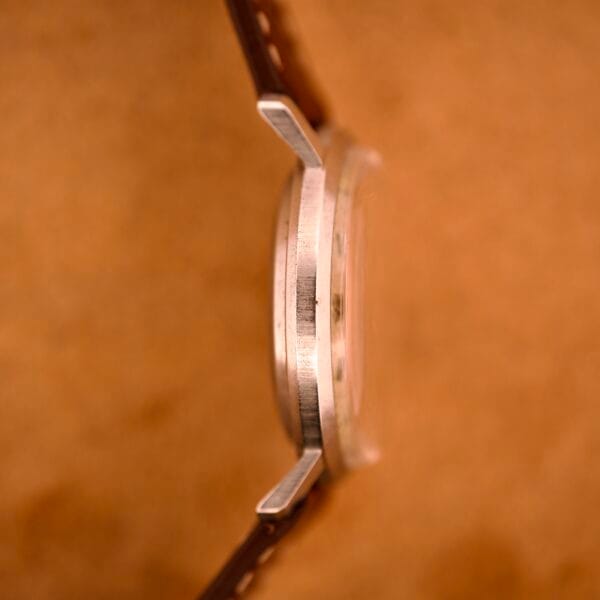 A close-up side view of the Bulova 30 Jewels Automatic Cal. 10COAC with a brown strap and Waffle Dial, featuring a stainless steel case, set against a blurred brown background.