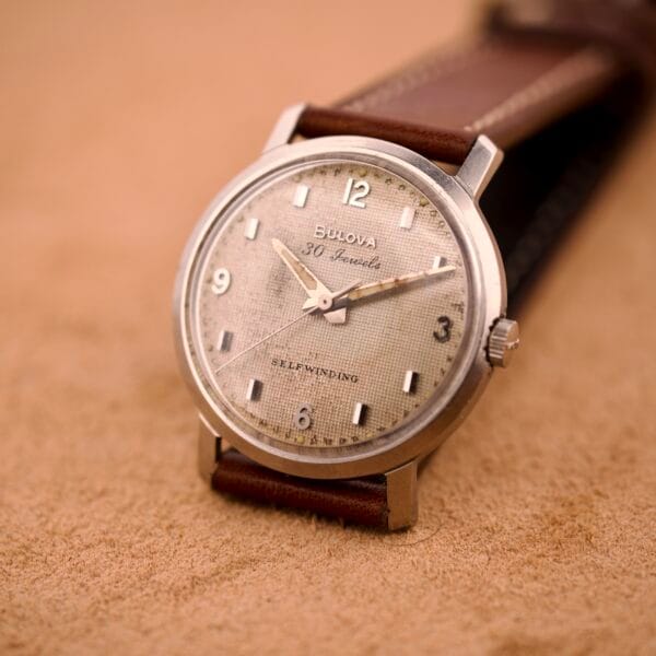 A Bulova 30 Jewels Automatic Cal. 10COAC wristwatch with a waffle dial and stainless steel case rests on a tan suede surface.