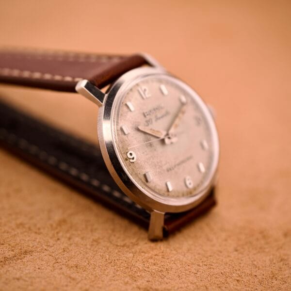 Close-up of a Bulova 30 Jewels Automatic Cal. 10COAC wristwatch with waffle dial and stainless steel case, featuring a brown leather strap, resting on a tan textured surface.