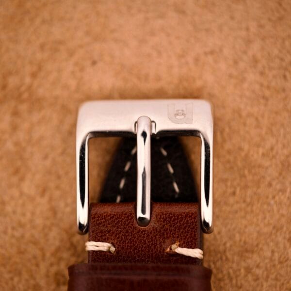 Close-up of the Bulova 30 Jewels Automatic Cal. 10COAC Waffle Dial's silver buckle on a brown leather strap, displayed against a tan textured background.