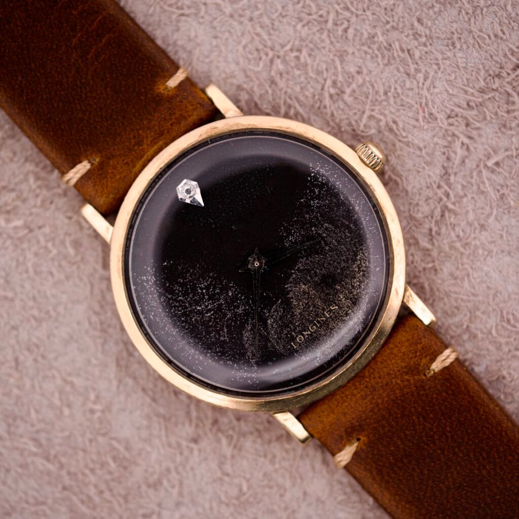 A 1970s Longines manual wind wristwatch with a black dial featuring diamond markers and a brown leather strap rests on a beige textured surface.