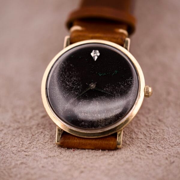 A 1970s Longines Manual Wind watch with a black dial and diamond marker rests on beige fabric, exuding vintage charm.