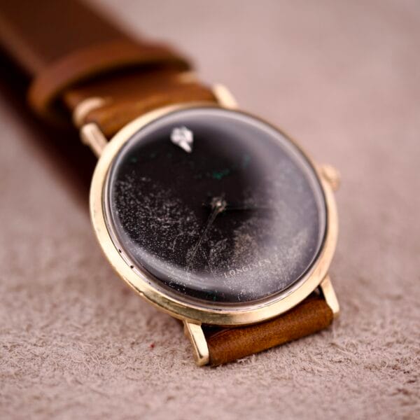 A 1970s Longines Manual Wind watch with a black dial, diamond marker, and a brown leather strap rests on a beige surface.