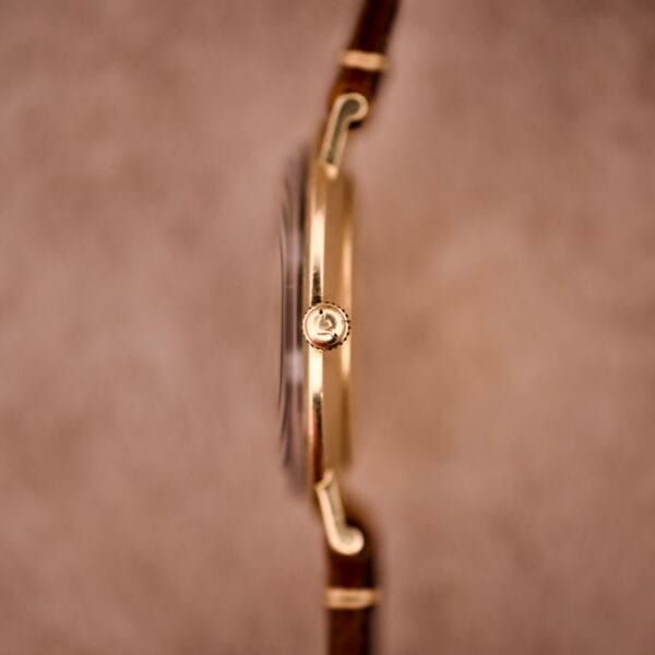 Side view of a 1970s Longines Manual Wind wristwatch with a black dial and diamond marker, featuring a gold case and brown leather strap, set against a blurred beige background.