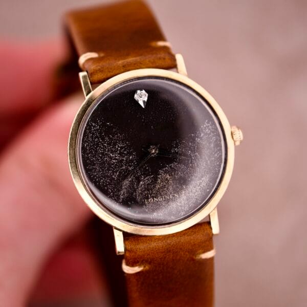 A 1970s Longines manual wind watch with a black dial featuring diamond markers and a brown leather strap; vintage charm with a cracked, scratched glass face.