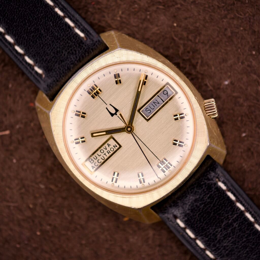 A Bulova Accutron Turning Fork Cal. 2182 Day-Date Cushion Case wristwatch with a gold finish, black leather strap, and iconic tuning fork movement, featuring a dial that shows both day and date.